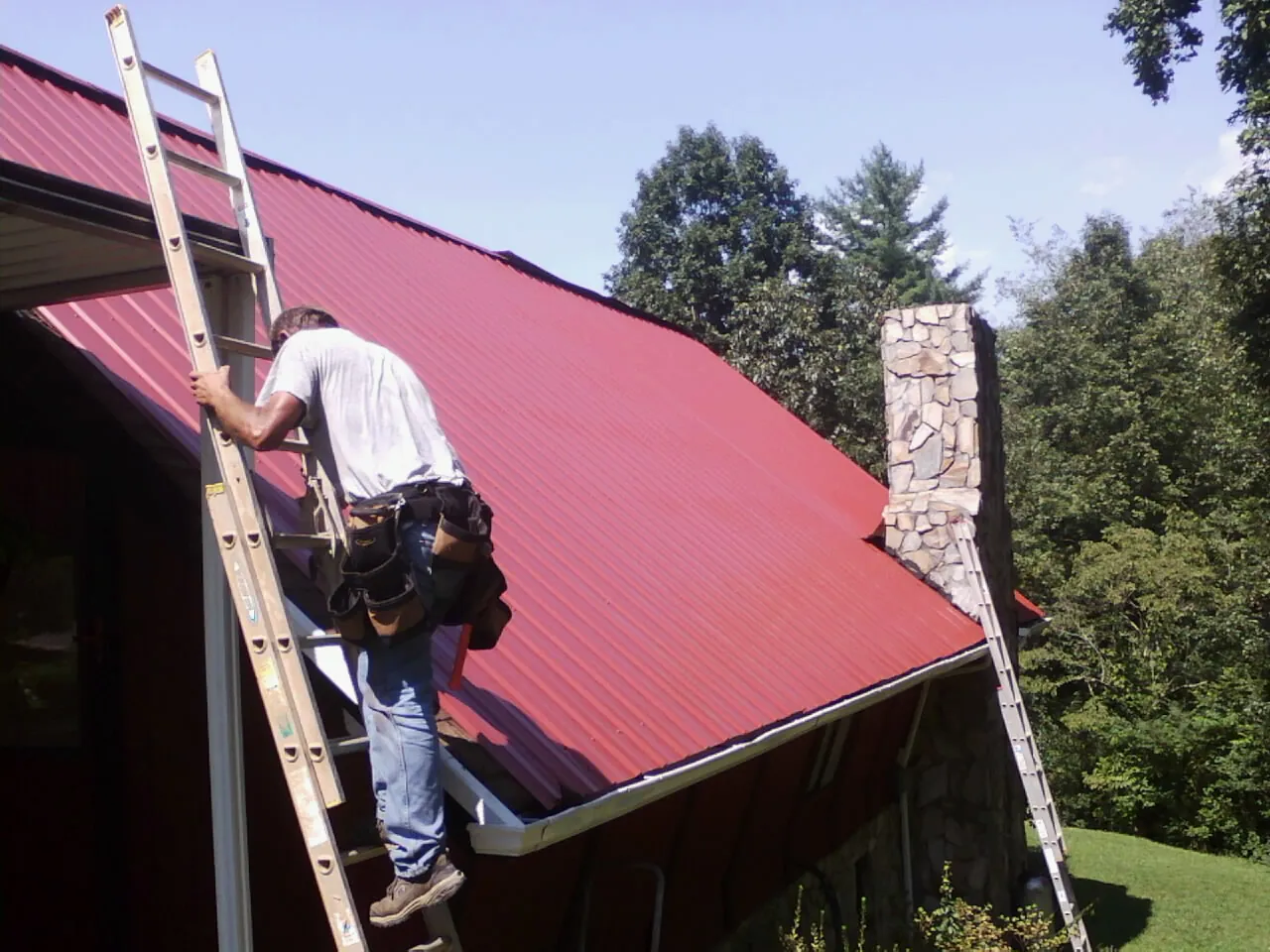 Roofer on a ladder beside a red metal roof and stone chimney — Soffit & Fascia Repair by Roofing Brothers in Cranesville